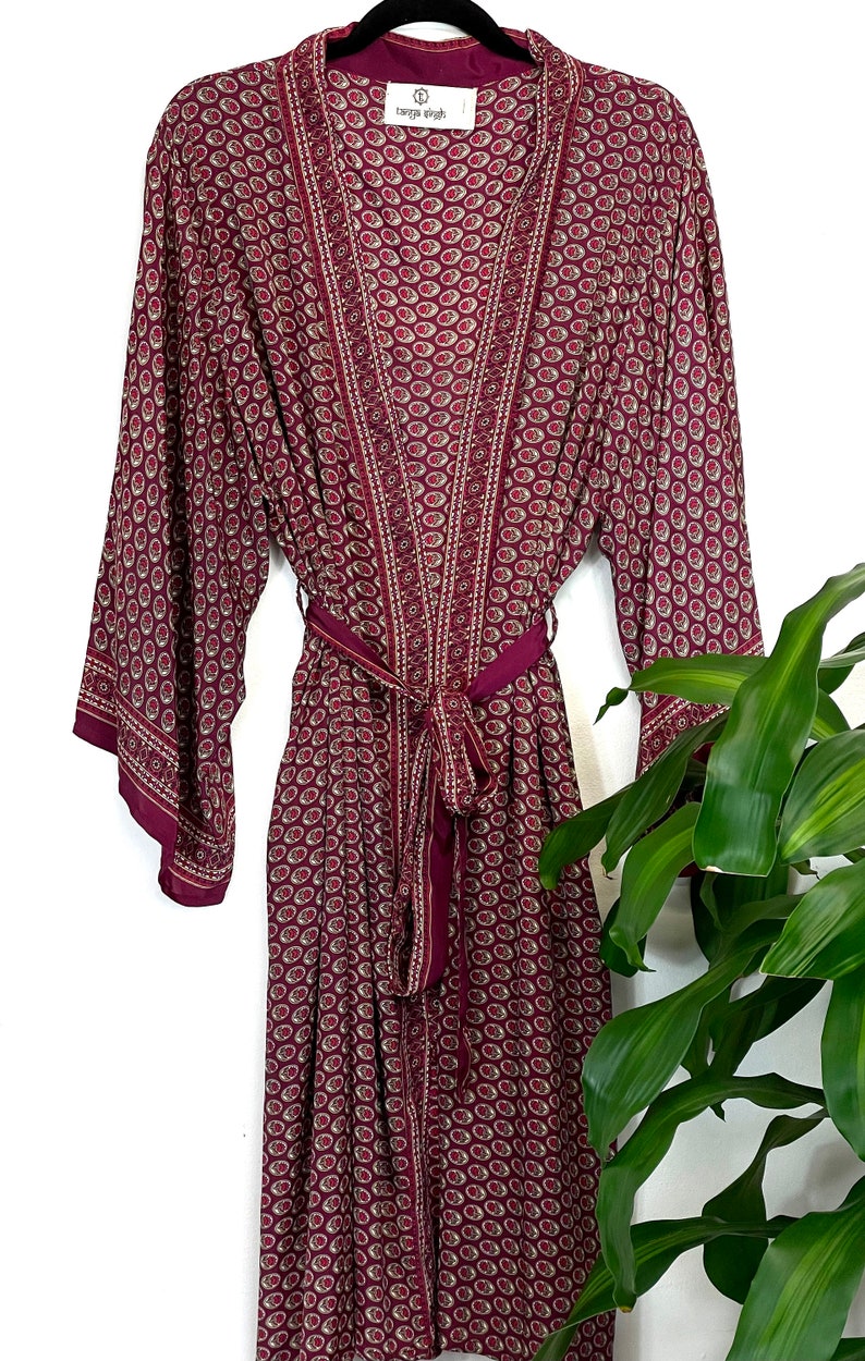 May include: A long, burgundy silk robe with a paisley print and a matching tie belt. The robe has a V-neck and long sleeves. The label on the robe reads "Tanga Nights".