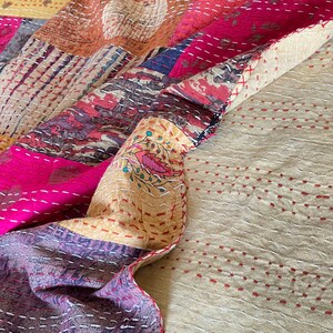 May include: A patchwork quilt featuring a mix of colours and patterns, including pink, yellow, brown, and blue. The quilt is made from various fabrics sewn together, with a hand-stitched appearance. The quilt is folded in half, with the bottom half being a plain beige colour with red stitching.