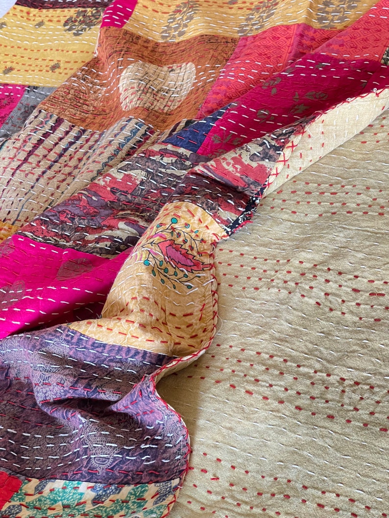 May include: A patchwork quilt featuring a mix of colours and patterns, including pink, yellow, brown, and blue. The quilt is made from various fabrics sewn together, with a hand-stitched appearance. The quilt is folded in half, with the bottom half being a plain beige colour with red stitching.