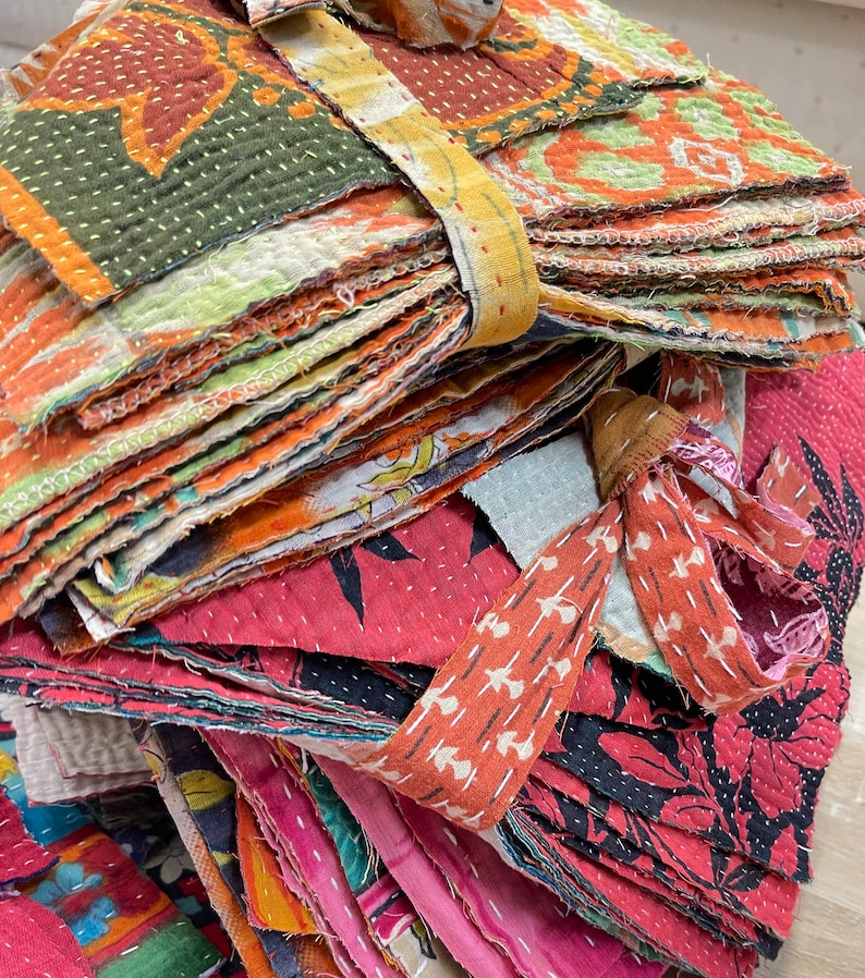 May include: A stack of colorful fabric squares, some with intricate stitching, tied together with a brown fabric ribbon. The fabric squares are a mix of red, orange, green, and blue.