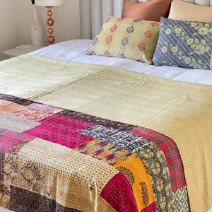 May include: A patchwork kantha quilt in shades of yellow, pink, red, blue, and brown. The quilt is made of various fabrics sewn together and features intricate hand stitching. The quilt is draped over a white bed with decorative pillows.