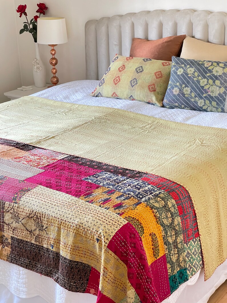 May include: A patchwork kantha quilt in shades of yellow, pink, red, blue, and brown. The quilt is made of various fabrics sewn together and features intricate hand stitching. The quilt is draped over a white bed with decorative pillows.