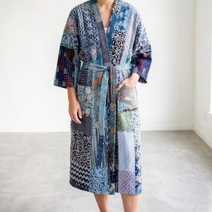 Pode incluir: Um roupão até o joelho com um design de patchwork composto por vários padrões de tecido em tons de azul, branco e vermelho escuro. A peça inclui mangas três quartos, um cinto de tecido a condizer na cintura e dois bolsos frontais.