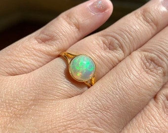May include: A gold ring with a round, white opal gemstone. The opal has a green and blue iridescent sheen.