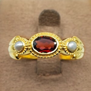 Könnte beinhalten: Ein goldener Ring mit einem roten Edelstein und zwei weißen Perlen. Der Ring hat ein detailliertes Design mit einem Perlenrand um den Edelstein.