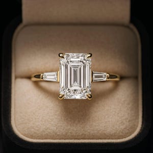 May include: A gold ring featuring a large emerald-cut center stone flanked by two smaller baguette-cut stones on each side. The ring is set in a polished yellow gold band, showcasing a classic three-stone design with clear, faceted gemstones.