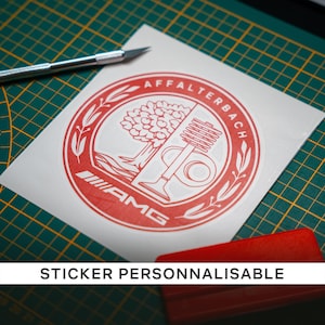 Peut inclure: Un autocollant rouge et blanc avec le logo AMG et le texte "AFFALTERBACH" dans un design circulaire. L'autocollant est sur un fond blanc, avec un cutter et un applicateur rouge visibles. Le texte "STICKER PERSONNALISABLE" est également présent.