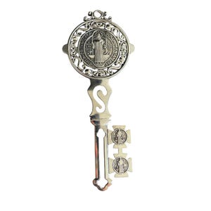 May include: Silver decorative key with an intricate design. The key has a circular top with a religious figure and text, an 'S' shaped handle, and two cross-shaped details at the bottom. The key is likely made of metal.