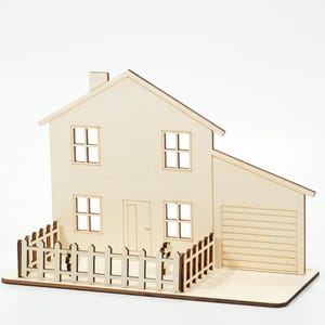 May include: A light-colored wooden model of a two-story house featuring a peaked roof, a chimney, four square windows, a central door, and an attached garage. A small wooden picket fence surrounds the front yard area of the structure.