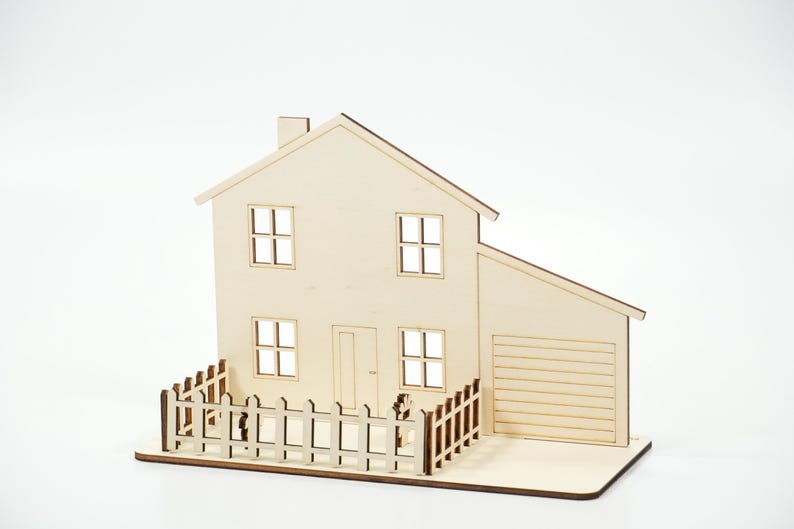 May include: A light-colored wooden model of a two-story house featuring a peaked roof, a chimney, four square windows, a central door, and an attached garage. A small wooden picket fence surrounds the front yard area of the structure.