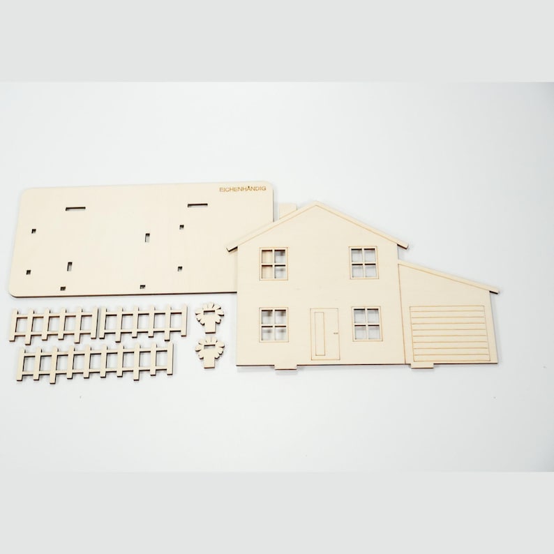 May include: A light-colored wooden model house kit featuring a two-story structure with a garage, four windows, a door, two fence sections, and two small tree cutouts. The flat pieces include a base plate with the brand name EICHENN&Auml;NDIG printed.