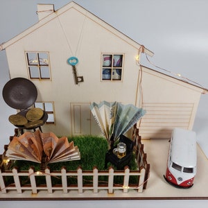 May include: A wooden house model featuring a miniature garden with a picket fence, a toy van, a grill with gold coins, folded euro banknotes, a small chair with a beer mug, a decorative key hanging on the wall, and string lights wrapped around the structure.