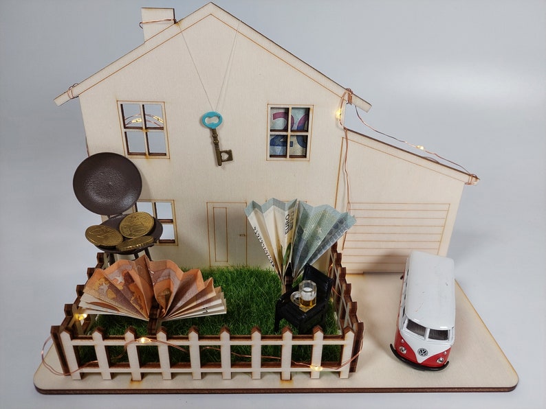 May include: A wooden house model featuring a miniature garden with a picket fence, a toy van, a grill with gold coins, folded euro banknotes, a small chair with a beer mug, a decorative key hanging on the wall, and string lights wrapped around the structure.