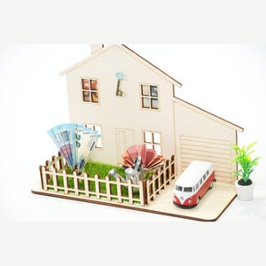 May include: A wooden house model featuring a fenced front yard with artificial green grass, a miniature metal watering can, a red and white toy van, a small potted plant, and paper currency folded into fans placed inside the fenced area.