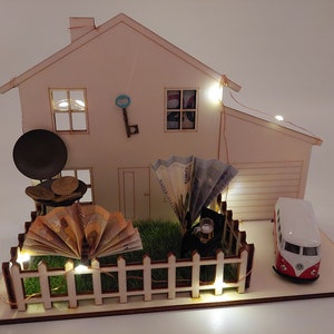 May include: A wooden house model featuring a white picket fence, green artificial grass, a miniature grill with coins, a fan-folded banknote, a small toy van, a hanging metal key, and warm LED string lights illuminating the structure.