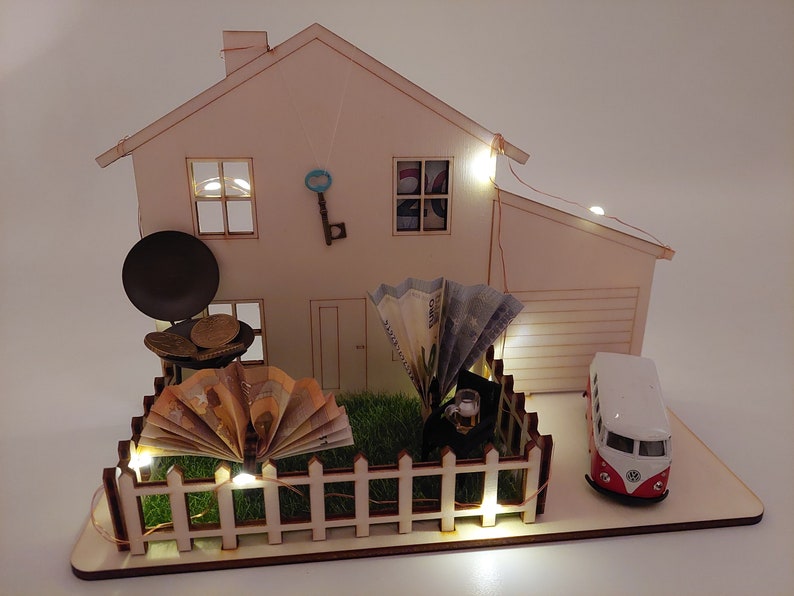 May include: A wooden house model featuring a white picket fence, green artificial grass, a miniature grill with coins, a fan-folded banknote, a small toy van, a hanging metal key, and warm LED string lights illuminating the structure.
