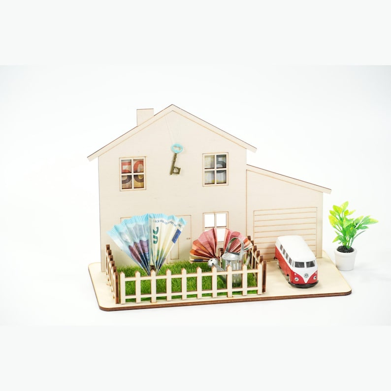 May include: A wooden house model featuring a white picket fence, green artificial grass, and a miniature red and white van. Euro banknotes are folded into fans in the yard, with a metal watering can, a decorative key, and a small potted plant.