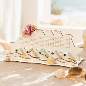 May include: A light-colored wooden cruise ship cutout featuring multiple rectangular window engravings and circular holes for rolled currency. The base is decorated with wave patterns and small seashells, with a fan-folded bill on top.