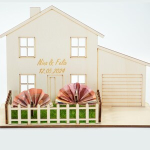 May include: A light-colored wooden house model featuring a garage, a white picket fence, and green artificial grass. The front of the house is engraved with the text Nina & Felix and the date 12.05.2024. Two folded paper currency fans are placed inside the fence.