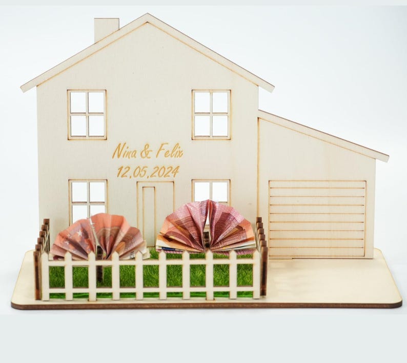 May include: A light-colored wooden house model featuring a garage, a white picket fence, and green artificial grass. The front of the house is engraved with the text Nina & Felix and the date 12.05.2024. Two folded paper currency fans are placed inside the fence.