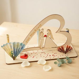 May include: A light-colored wooden heart-shaped display piece featuring a cutout of a couple standing together. The text "zur Hochzeit" is engraved on the heart. The base includes folded paper currency, a small red heart, and decorative petals.