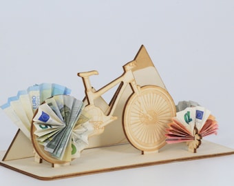 May include: A wooden bicycle sculpture with euro banknotes arranged to resemble the wheels. The sculpture is on a wooden base with a mountain-shaped element. The euro bills are in various denominations, with blue, green, and orange hues.