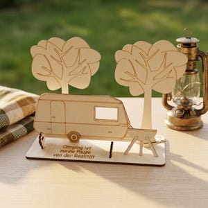 May include: A light-colored wooden decorative display featuring a caravan, two trees, and a small grill on a rectangular base. The base is engraved with the German text 'Camping ist meine Pause von der Realität' in a simple, dark sans-serif font.