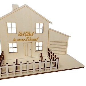 May include: A light-colored wooden house model featuring a two-story structure with a garage, four windows, a front door, and a picket fence. The front of the house is engraved with the German text "Viel Gl&uuml;ck im neuen Zuhause!" in a script font.