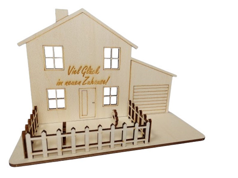 May include: A light-colored wooden house model featuring a two-story structure with a garage, four windows, a front door, and a picket fence. The front of the house is engraved with the German text "Viel Gl&uuml;ck im neuen Zuhause!" in a script font.