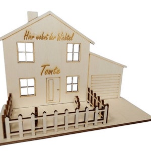 May include: A light-colored wooden house model featuring a chimney, four windows, a door, and an attached garage. The text "Hier wohnt der Wichtel" and "Tomte" are engraved on the front. A small picket fence surrounds the front yard area.