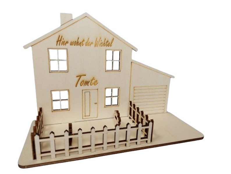 May include: A light-colored wooden house model featuring a chimney, four windows, a door, and an attached garage. The text "Hier wohnt der Wichtel" and "Tomte" are engraved on the front. A small picket fence surrounds the front yard area.