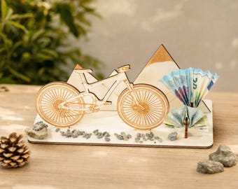 May include: A laser-cut wooden mountain bike silhouette mounted on a rectangular base with two triangular mountain shapes in the background. The display features small grey stones scattered on the base and folded paper currency fan accents.