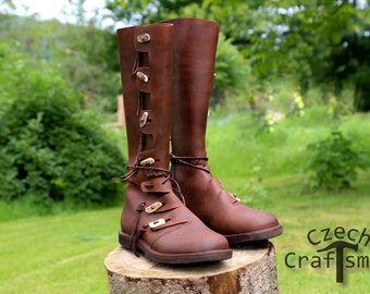 Könnte beinhalten: Ein Paar braune, kniehohe Lederstiefel mit dekorativen Schnallen und Schnürsenkeln. Die Stiefel stehen auf einem Baumstumpf in einem grasbewachsenen Bereich.