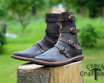 May include: A pair of brown leather boots with multiple buckles. The boots have a pointed toe and a thick sole. The boots are sitting on a tree stump.