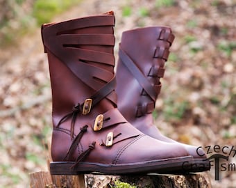 Könnte beinhalten: Ein Paar braune Lederstiefel mit dekorativen Riemen und Schnallen. Die Stiefel haben eine spitze Zehenpartie und einen niedrigen Absatz. Die Stiefel stehen auf einer Holzoberfläche.