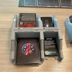 May include: A light grey 3D-printed plastic card organiser for board games with four distinct slots. The organiser features embossed text labels including HIVE HAZARDS, DISCARD, HIVEMIND, and NATURE, holding various game cards with printed text.