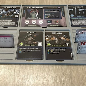 May include: A grey plastic board game organizer tray holding several rectangular game cards. The cards feature character portraits, weapon stats, and equipment details including M41A Pulse Rifle, Pvt. Jenette Vasquez, M4 Helmet, and Body Armor.