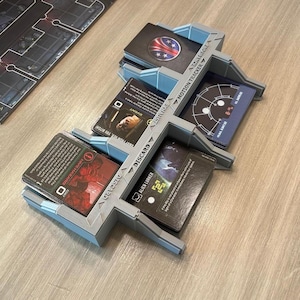 Puede incluir: Un organizador de cartas para juegos de mesa hecho de plástico gris claro con cuatro ranuras distintas etiquetadas con texto que incluye Motion Tracker, Exhaust y Discard. El organizador contiene varias cartas de juego con ilustraciones gráficas y texto, incluyendo Hive Hazard y Alien Lurker, dispuestas en un diseño estructurado.