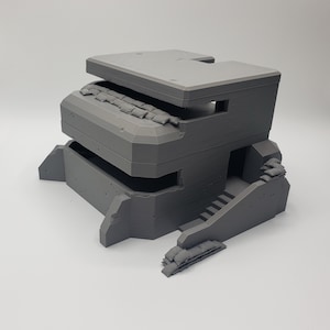 May include: A gray, 3D printed model of a concrete bunker with a roof, a doorway, and a set of stairs. The bunker has sandbags around the top and bottom.