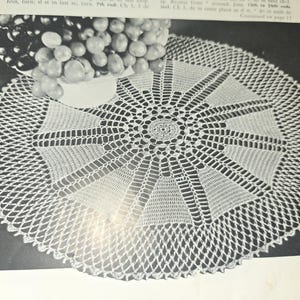 May include: A detailed, circular, white crocheted doily with a central floral design and a geometric pattern radiating outwards. The doily is placed on a dark surface, with a bowl of grapes in the upper left corner.