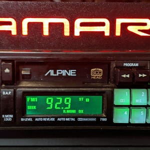 May include: A black car stereo with a green digital display showing "92.9" and the word "ALPINE" in white lettering. The stereo has a red "CAMARO" inscription above it.