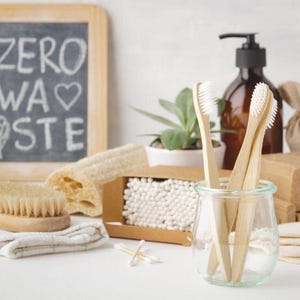 May include: A collection of zero-waste bathroom products, including bamboo toothbrushes, a wooden brush, soap, cotton swabs, and reusable cotton pads. A small chalkboard with the words "ZERO WASTE" is also present. The items are arranged on a white surface.