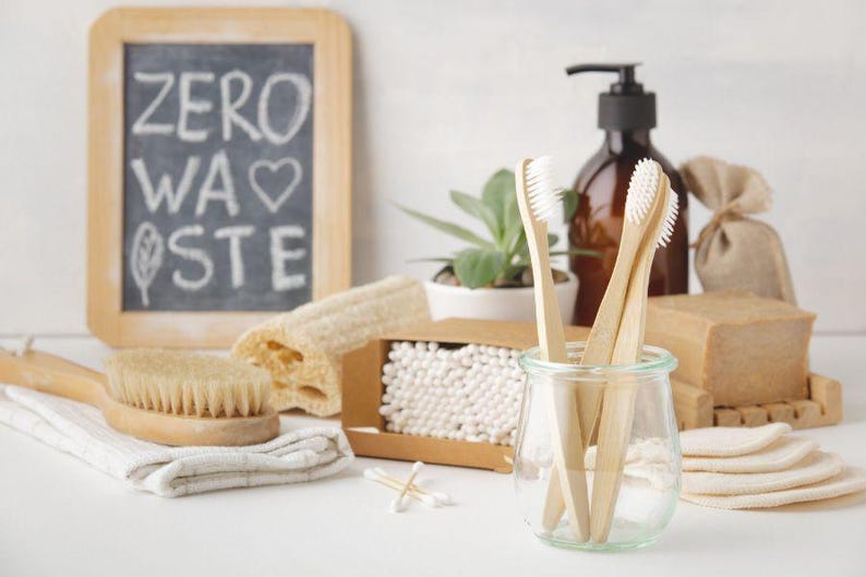 May include: A collection of zero-waste bathroom products, including bamboo toothbrushes, a wooden brush, soap, cotton swabs, and reusable cotton pads. A small chalkboard with the words "ZERO WASTE" is also present. The items are arranged on a white surface.