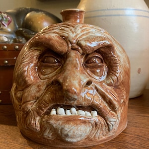 May include: A brown ceramic jug with a detailed, aged face design. The jug features a prominent nose, furrowed brow, and visible teeth. The jug has a small spout at the top and is likely a decorative or collectible item.