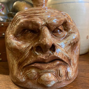 May include: A brown ceramic jug with a sculpted face, featuring furrowed brows, a prominent nose, and a downturned mouth. The jug has a small spout at the top and is glazed with a glossy finish. The overall design evokes a sense of age and character.