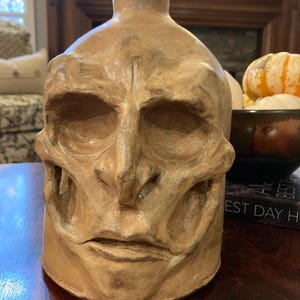 May include: A tan-colored ceramic vase with a sculpted human skull design. The vase has a narrow neck and a wide, rounded base. The skull features detailed eye sockets, a nose, and a mouth. The vase is likely handmade and suitable for home decor.