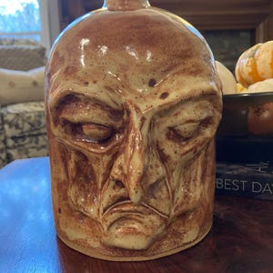 May include: A handcrafted ceramic jug with a detailed, sculpted face. The jug features a brown and tan color palette with a textured surface. The face has closed eyes, a prominent nose, and a downturned mouth, giving it a contemplative expression. The jug has a narrow neck.