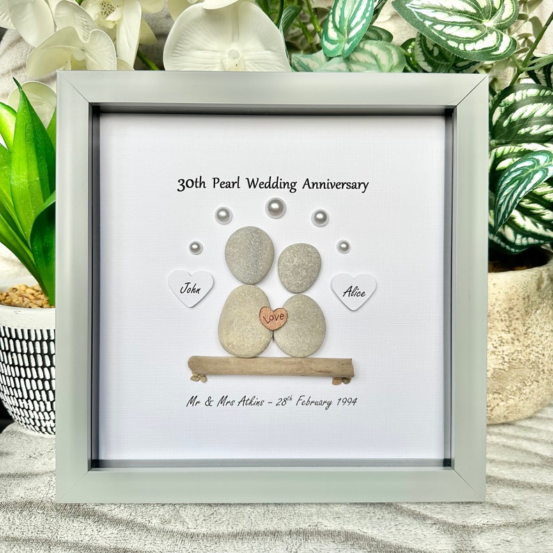 May include: A framed pebble art piece celebrating a 30th wedding anniversary. The art features two gray pebbles representing a couple, with the words "John" and "Alice" on white hearts. The text "30th Pearl Wedding Anniversary" and "Mr & Mrs Atkins - 28th February 1994" are also included.