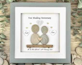 May include: A framed artwork for a first wedding anniversary. The artwork features two pebble figures, a wooden base, and the word "Love". The frame is gray, and the background is white with the text "First Wedding Anniversary".