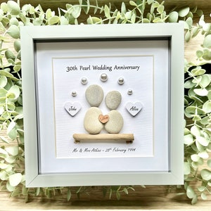 May include: A white framed picture with a 30th Pearl Wedding Anniversary message. The picture features two gray stones representing a couple, with a small wooden heart that says "Love" between them. The stones are sitting on a small piece of driftwood. The names "John" and "Alice" are written on small white hearts below the stones. The date "28th February 1994" is written below the driftwood.